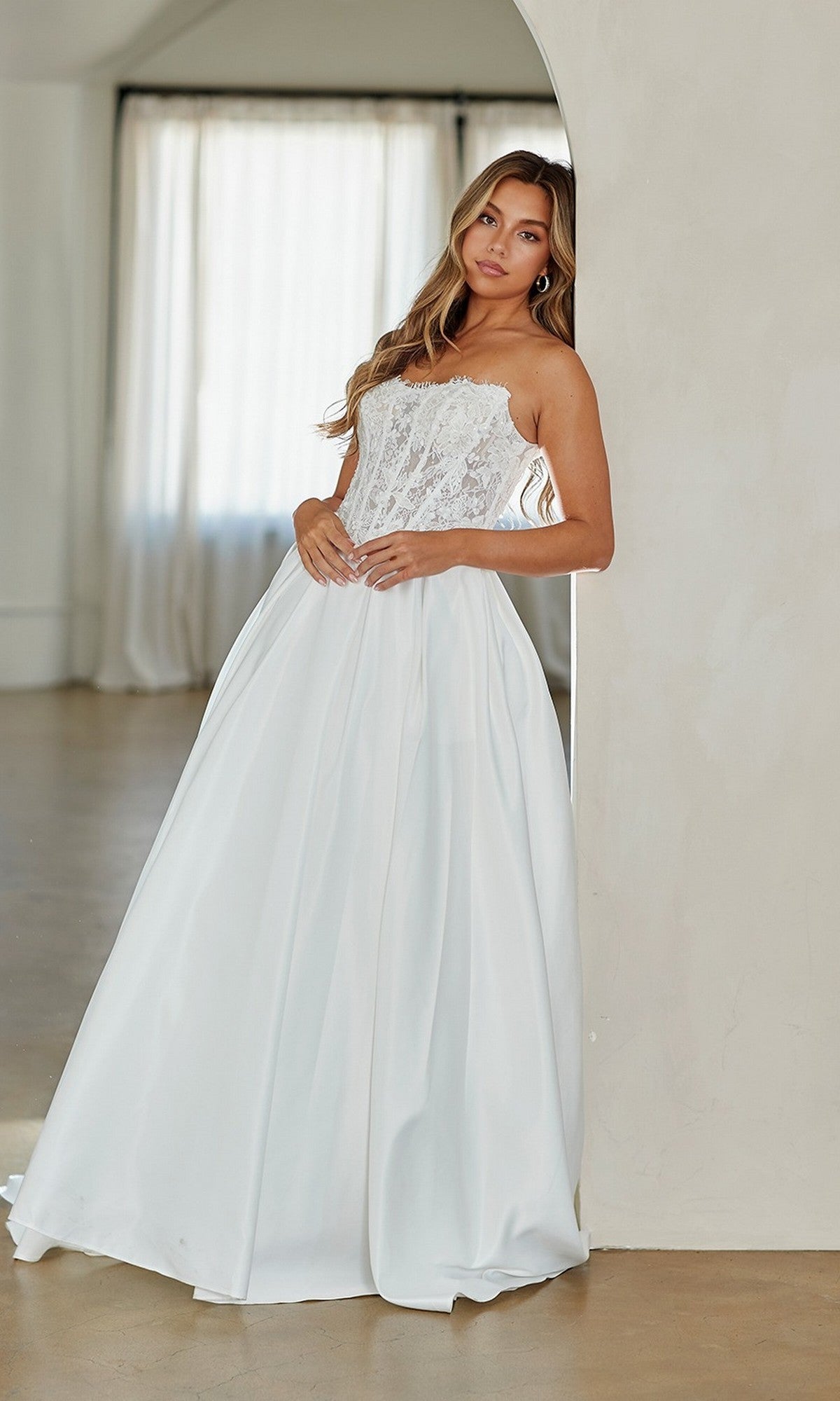 A woman in the Amelia Couture 2089 long prom dress, featuring a strapless white lace corset bodice and satin skirt, stands by an arched wall in a softly lit room.